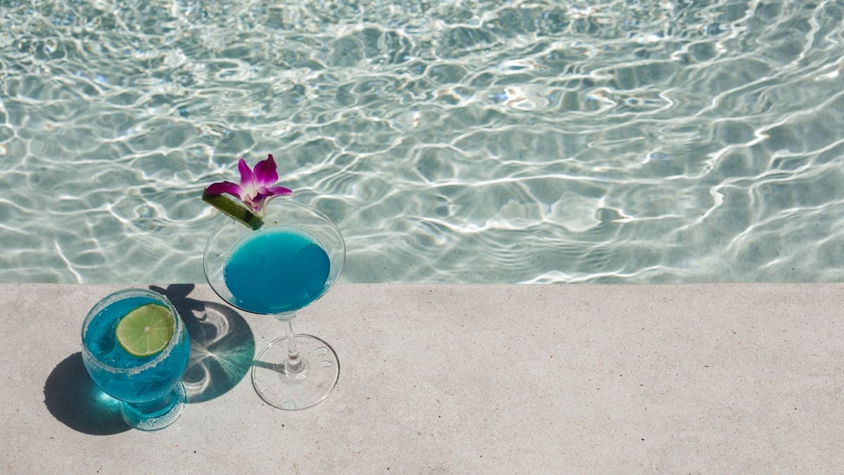 Two blue cocktails with lime and orchid by a sunlit poolside, perfect for a tropical vacation.